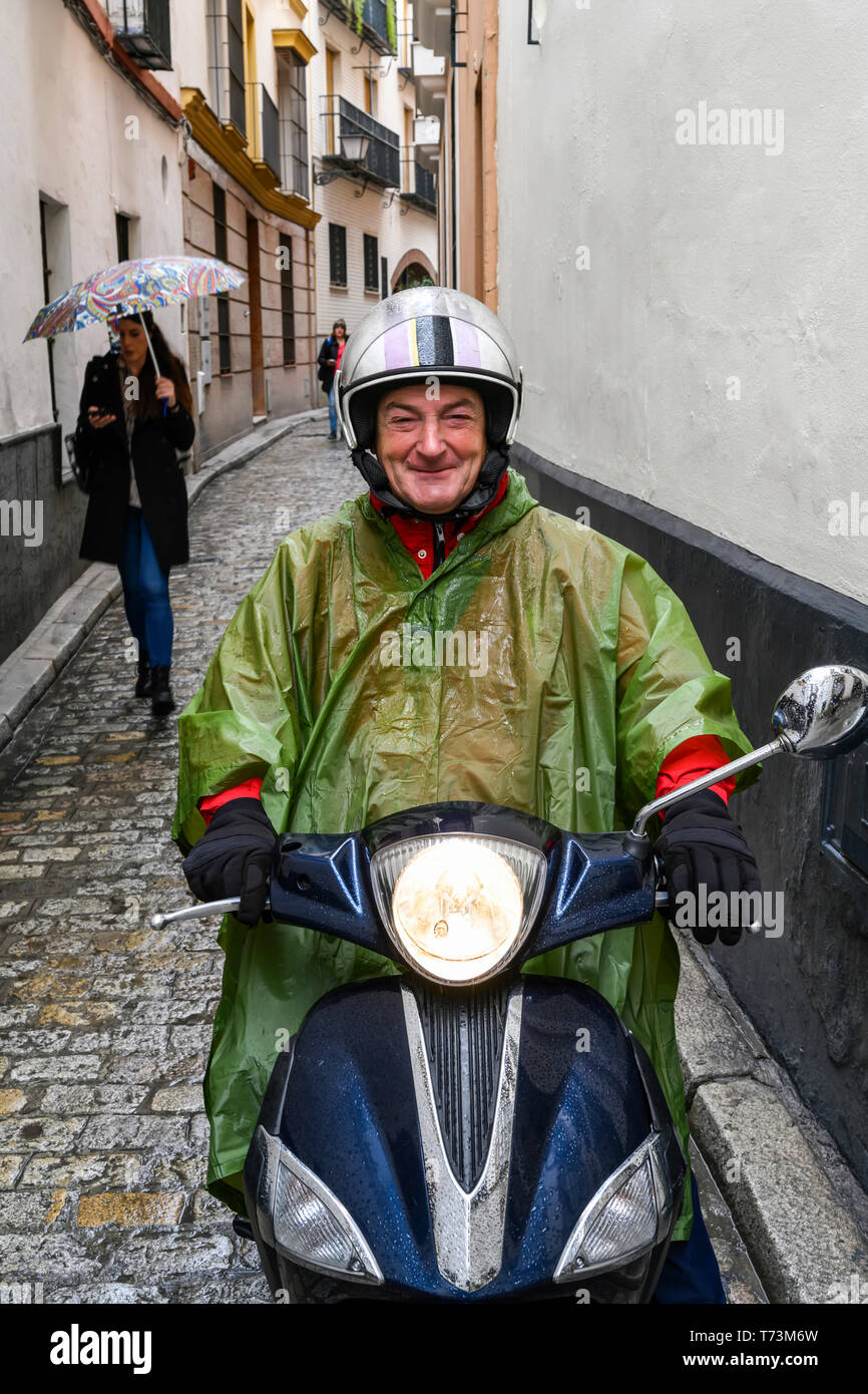 A man wearing a poncho and helmet sits on a motor scooter on a narrow street posing for the camera; Seville, Spain Stock Photo