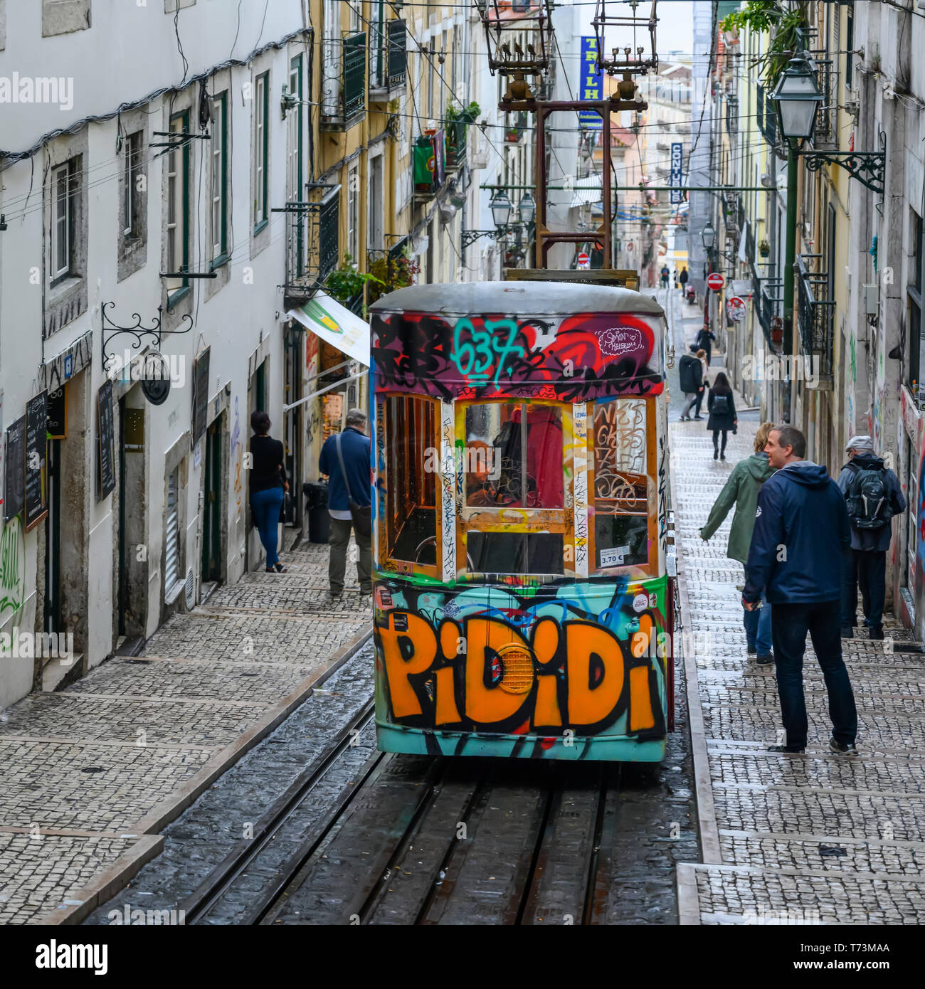 Street car with graffiti going downhill on a sloped street with pedestrians and residential buildings; Lisbon, Lisboa Region, Portugal Stock Photo