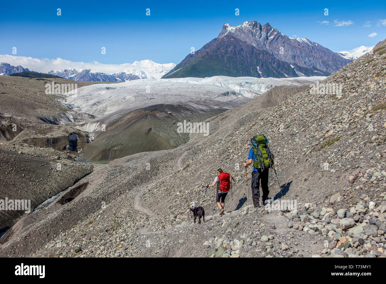 Couple and their dog backpacking on glacial morraine, heading towards the Root Glacier, with Donoho Peak and the Wrangell Mountains in the backgrou... Stock Photo