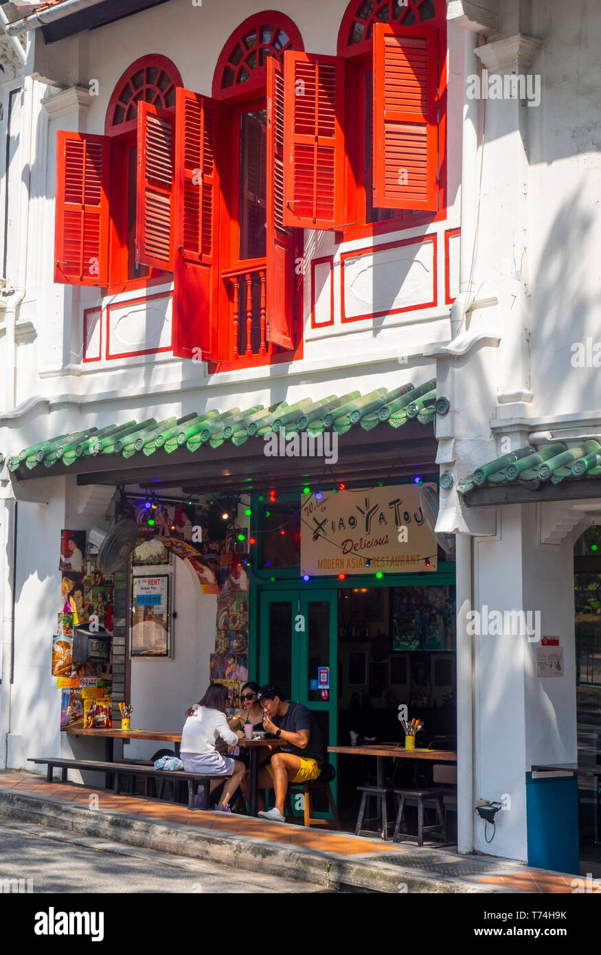 Locals eating alfresco outside a restaurant in a traditional shophouse with red window shutters. Stock Photo