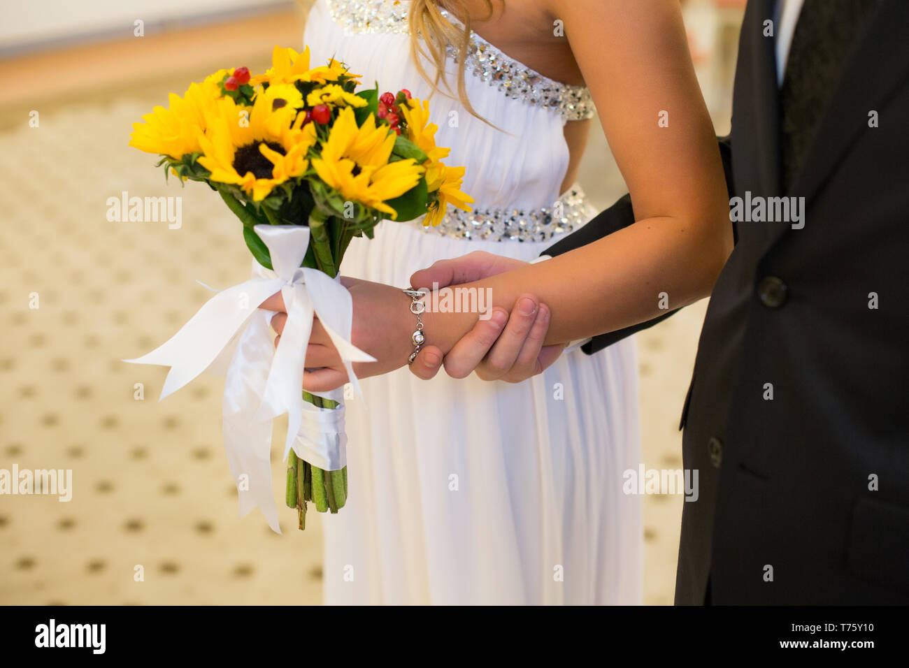 A miniature bouquet of flowers of a sunflower, supplemented with red hypericum berries, in the hands of an unrecognizable bride and groom Stock Photo