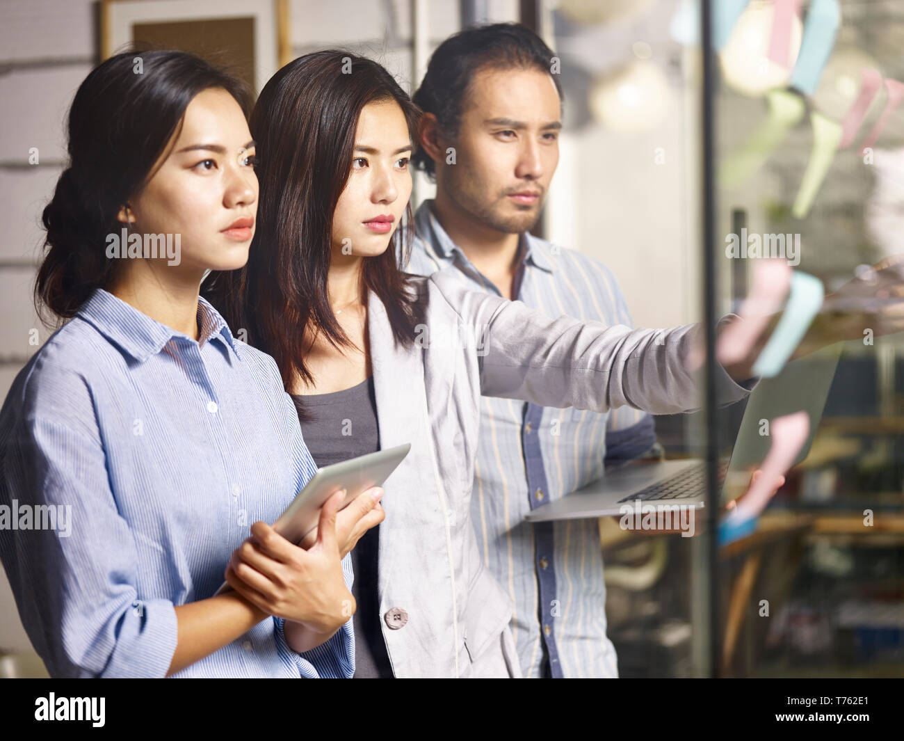 three young asian entrepreneurs teaming up analyzing business situation using laptop computer, digital tablet and adhesive notes in office, looking se Stock Photo