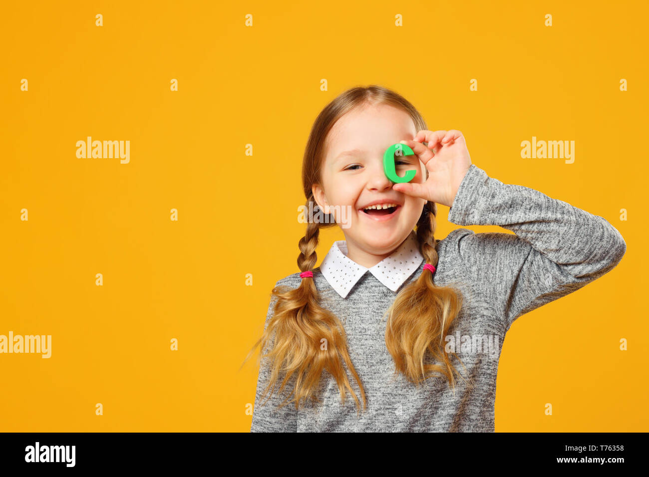 Portrait of a cheerful little child girl on a yellow background. Schoolgirl holds the letter C. The concept of education. Stock Photo