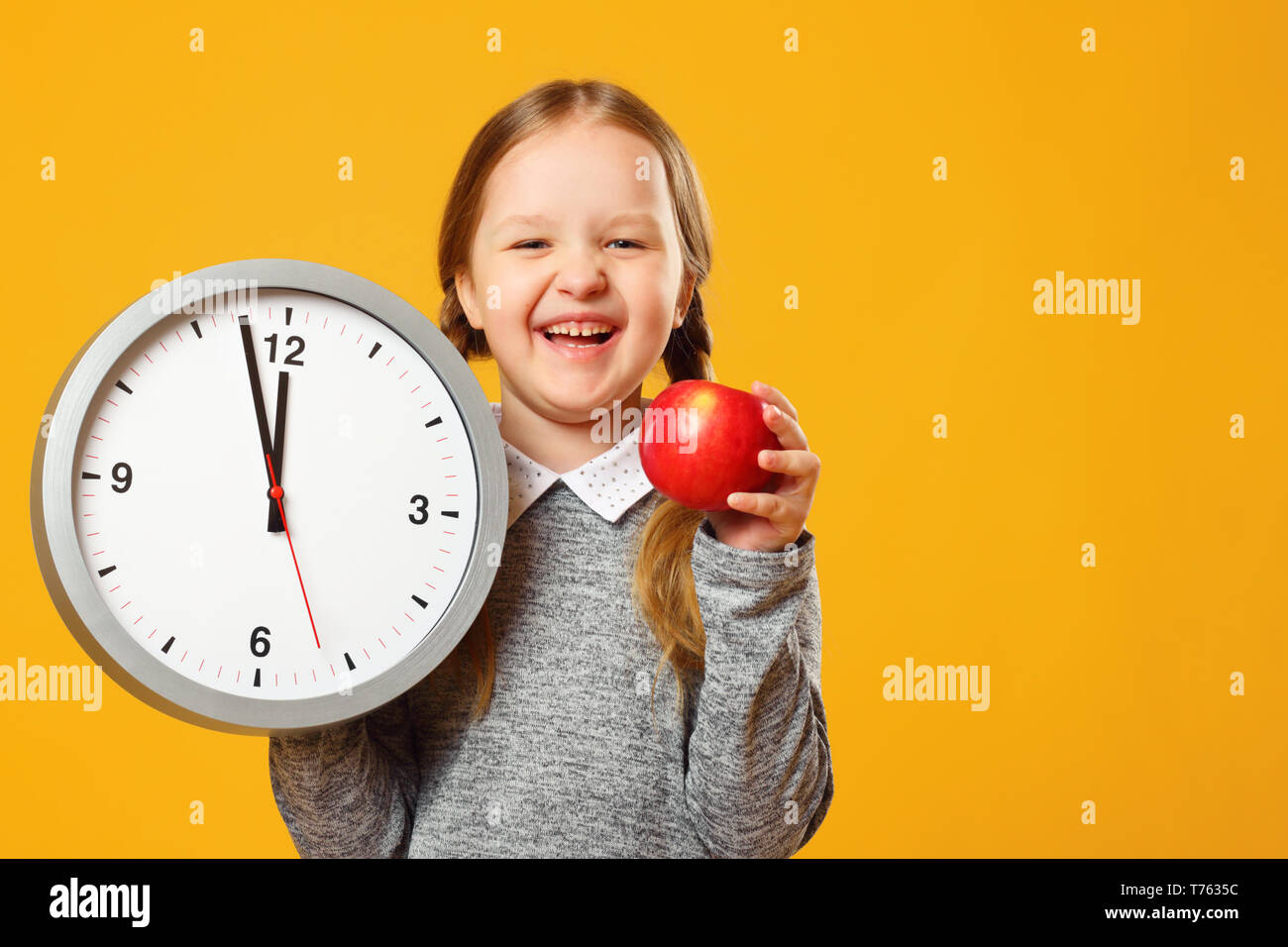 Little girl schoolgirl holds a big clock and a red apple on a yellow background. Break time and lunch. Stock Photo