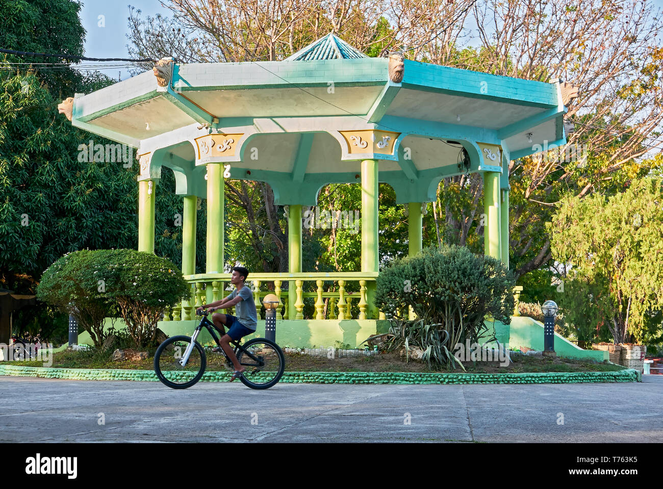 Leon, Iloilo, Philippines: Teenage boy riding a bike in front of a green colored pavilion at the town plaza park in summer time Stock Photo