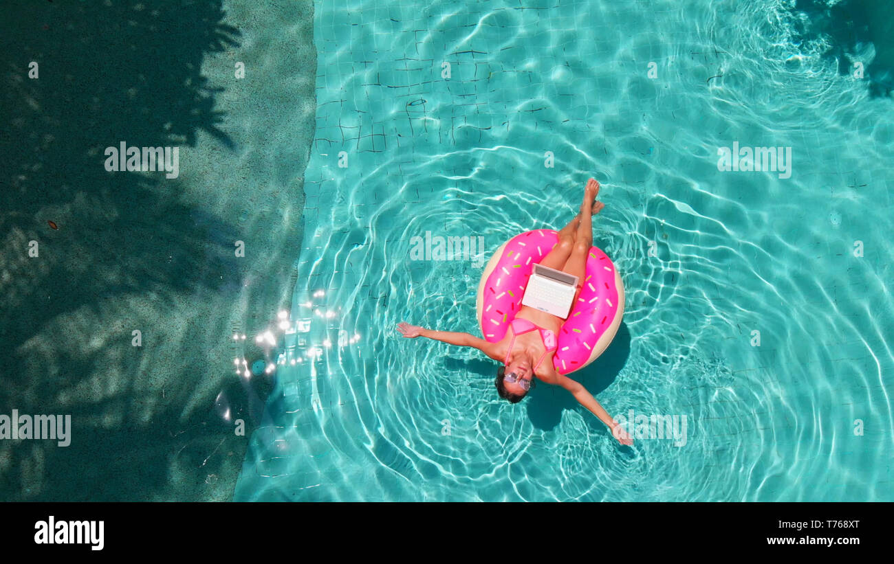 Beautiful young woman with a pink laptop on an inflatable ring in the water in the swimming pool. freelancing, business and travel concept. Aerial Stock Photo