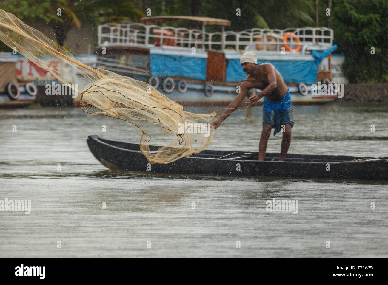Man stands on long dugout canoe in shallows of jungle river casting out a fishing net with native house boat in background Stock Photo