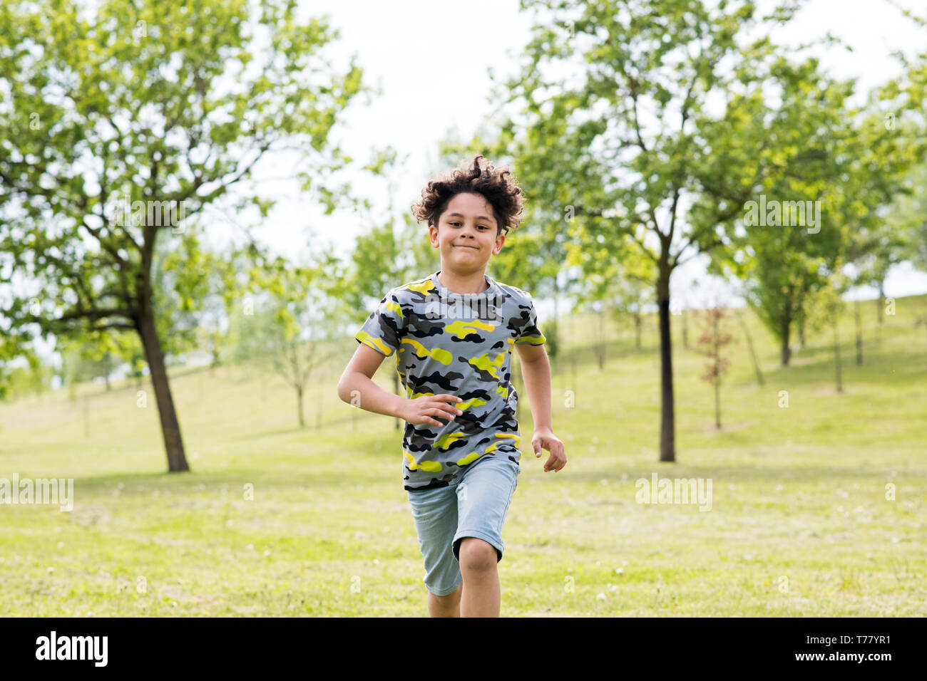 Happy fit young boy running towards the camera across the green grass in a spring garden with a big friendly smile Stock Photo