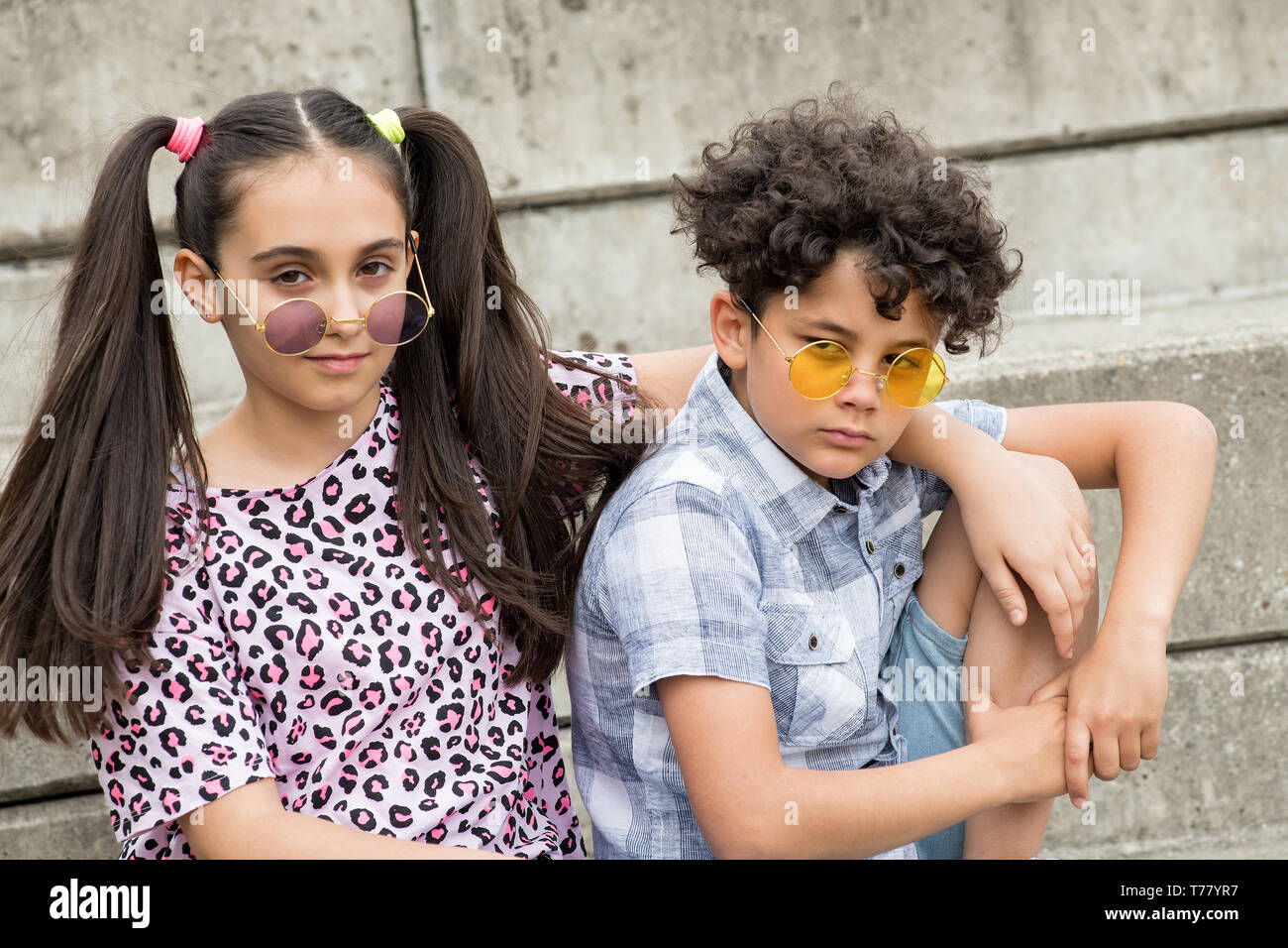 Trendy young brother and sister wearing modern colored circular sunglasses sitting arm in arm on outdoor steps looking at camera with serious expressi Stock Photo