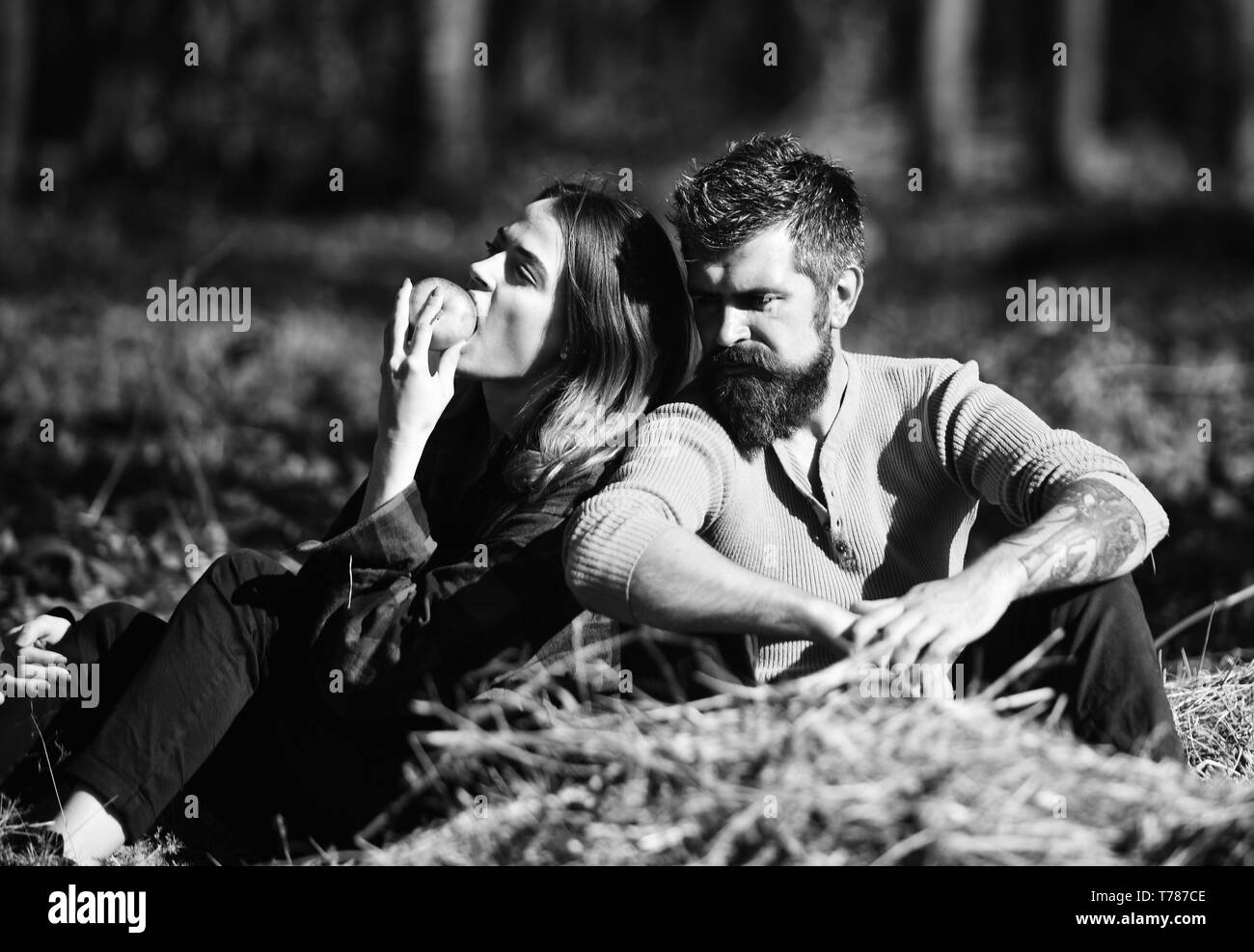 Relationship and fall time concept. Girl and bearded guy or lovers on a date sit on dry grass eating apple. Man and woman with serious faces on nature background. Couple in love spends time in park. Stock Photo