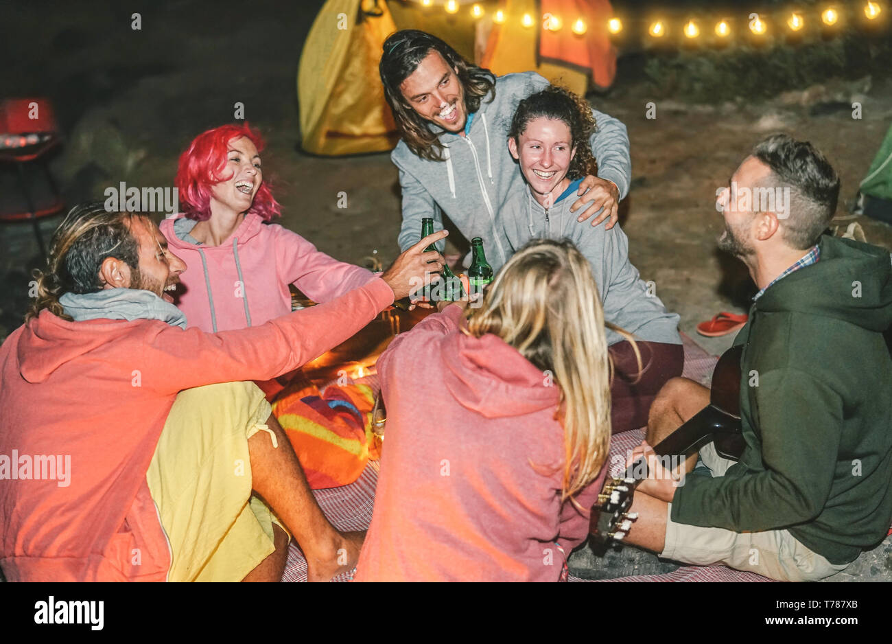 Group of friends having fun cheering with beers on the beach with tent at night - Happy young people playing guitar and laughing together Stock Photo