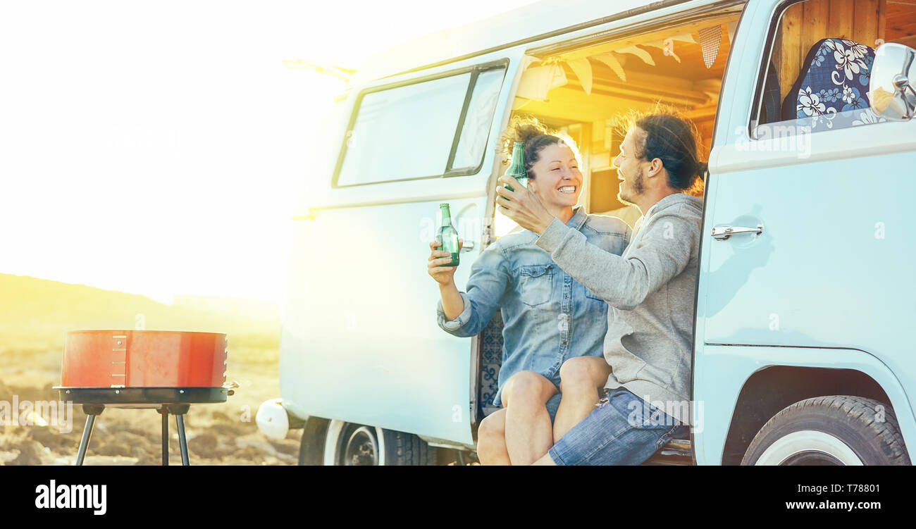 Travel couple having fun drinking beers sitting on their vintage camper mini van - Happy people with their pet enjoying barbecue at sunset Stock Photo