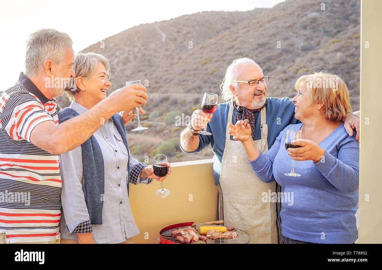 Happy senior friends having fun drinking red wine at barbecue in terrace - Mature people dining and laughing together on rooftop Stock Photo