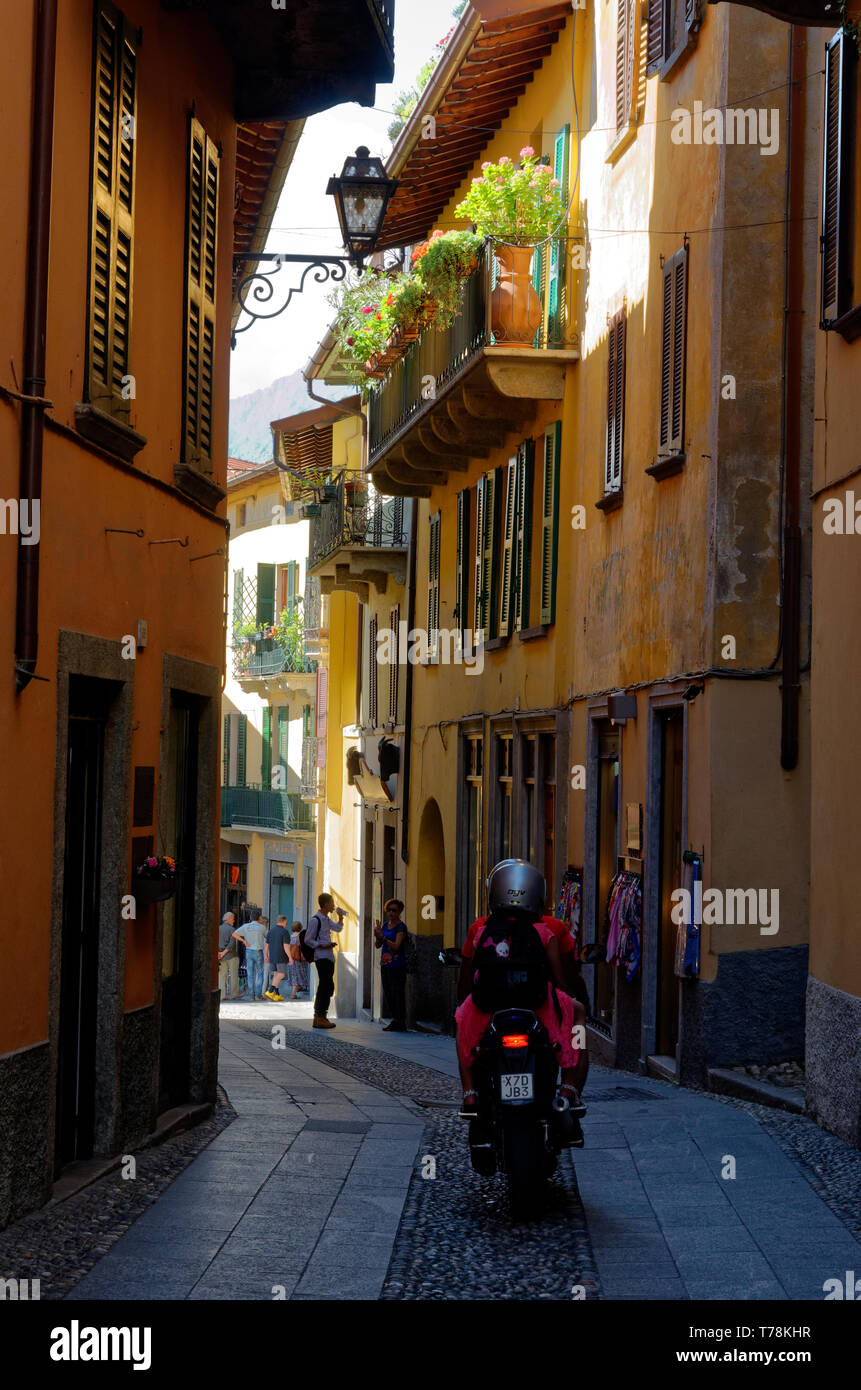 Motor scooter driving down the cobbles in the shade of a narrow lane in Bellagio, underneath flower boxes Stock Photo