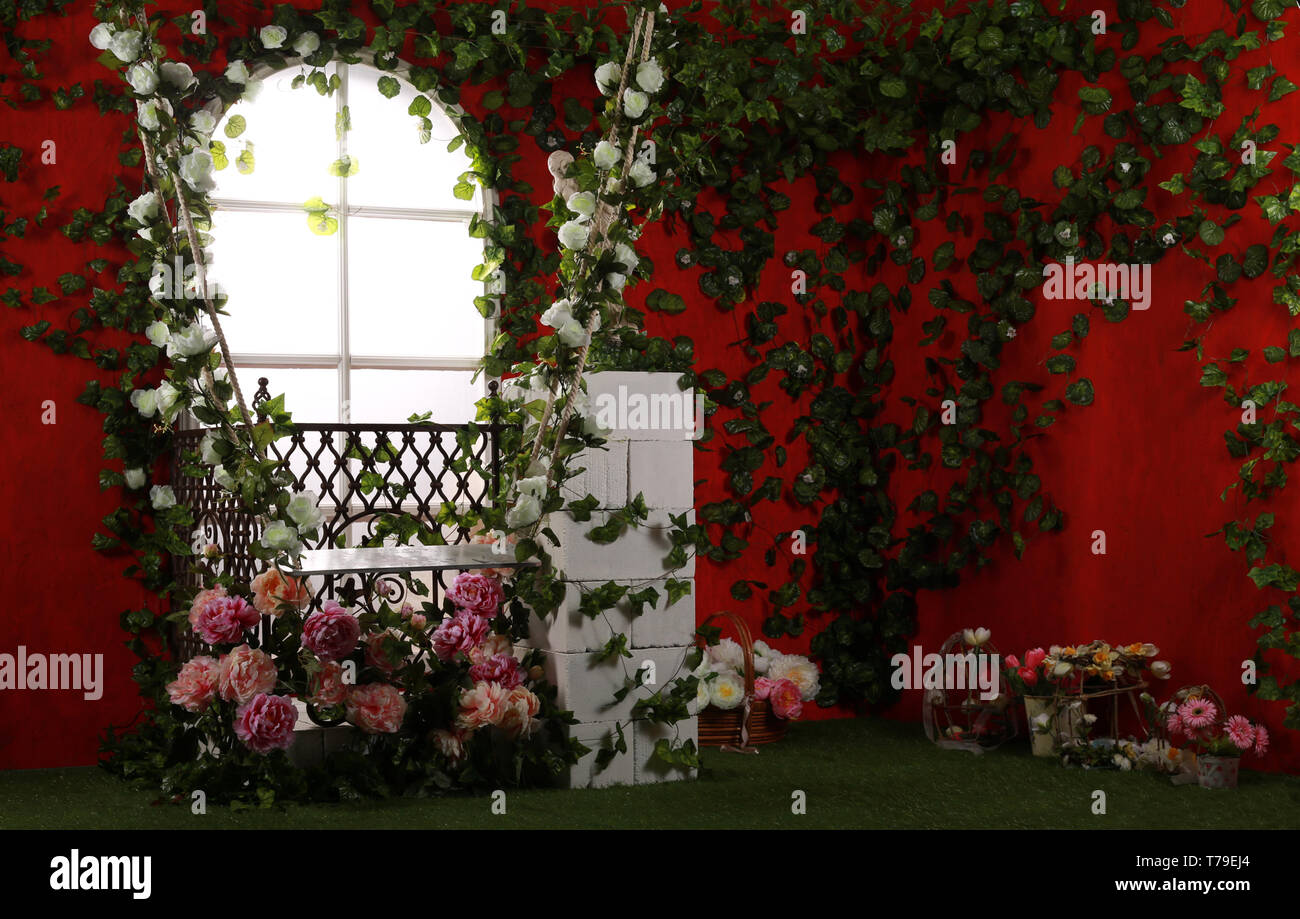 Photo zone, location with garden swings, decorated with flowers for the wedding. Against a bright red wall with blooming greenery. Stock Photo