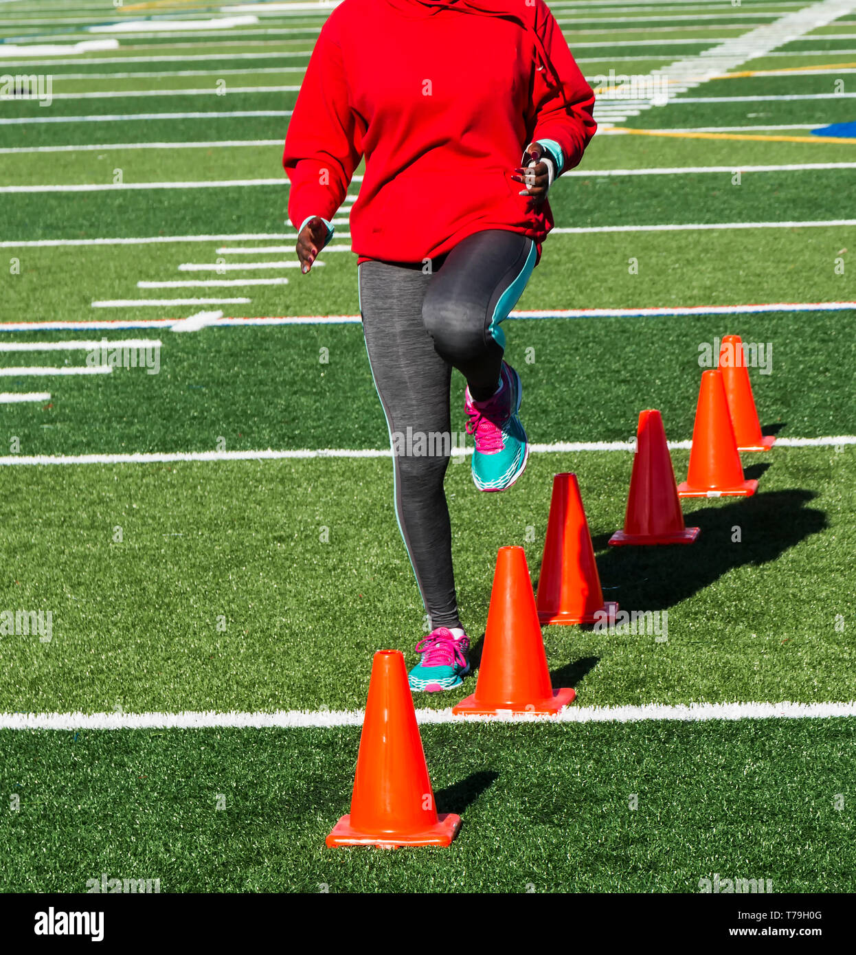 A high school female track and field runner steeping over orange cones performing speed drill on a green turf field. Stock Photo