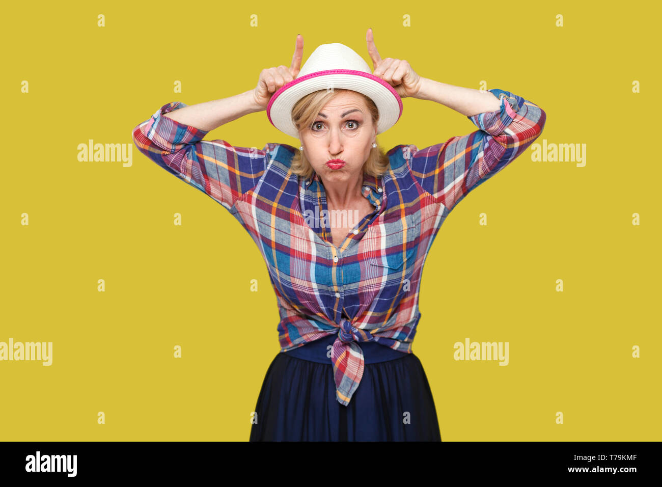 Portrait of crazy comic modern stylish mature woman in casual style with white hat standing with cow horns on head with hands and looking at camera. i Stock Photo