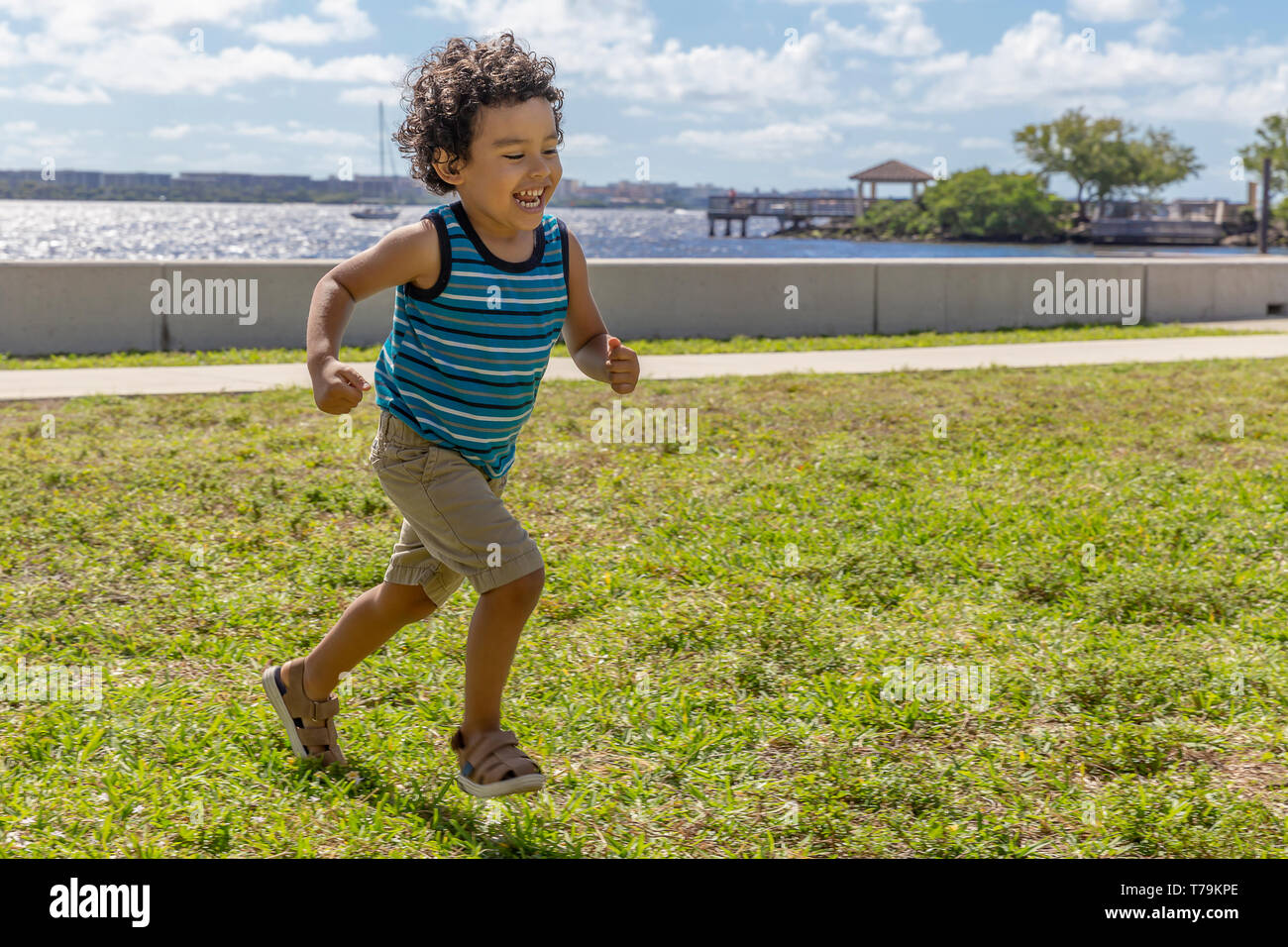 A young boy runs across the field while laughing out loud. The happy toddler is having a wonderful time running outdoors in the waterfront park. Stock Photo