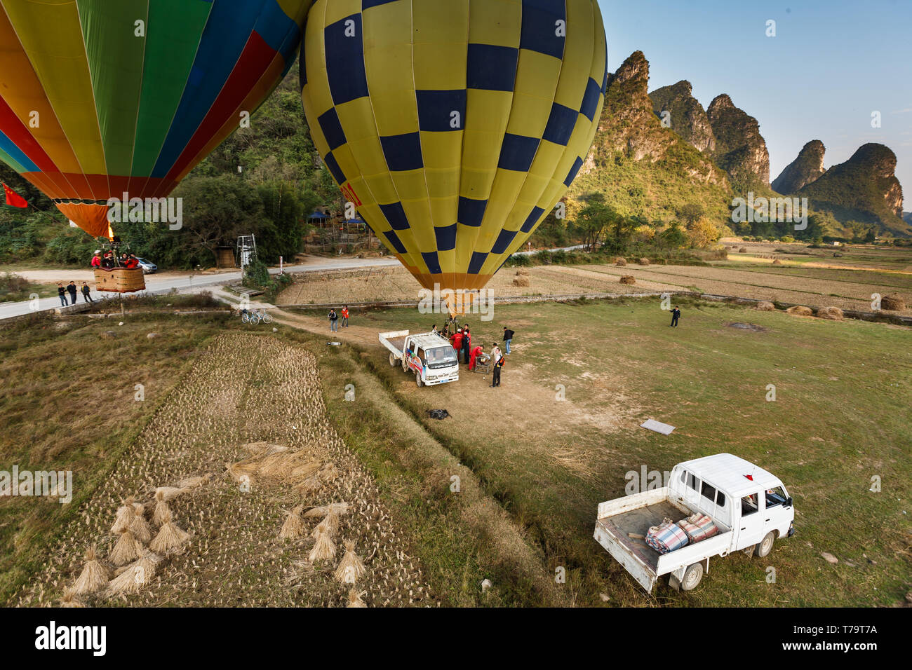 A high-angle shot captures two hot air balloons, one with a rainbow pattern and the other with a yellow and blue checkered design, hovering over a rural landscape with mountains in the background. Ground crews and trucks are positioned below the balloons, likely assisting with the landing or preparation for flight, amidst harvested fields and natural terrain. Stock Photo