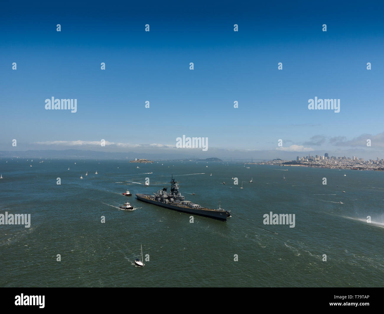 View of the side of a navy world war II battleship escorted by tugboats leaving San Francisco harbor with hundreds of sailboats dotting water beyond Stock Photo