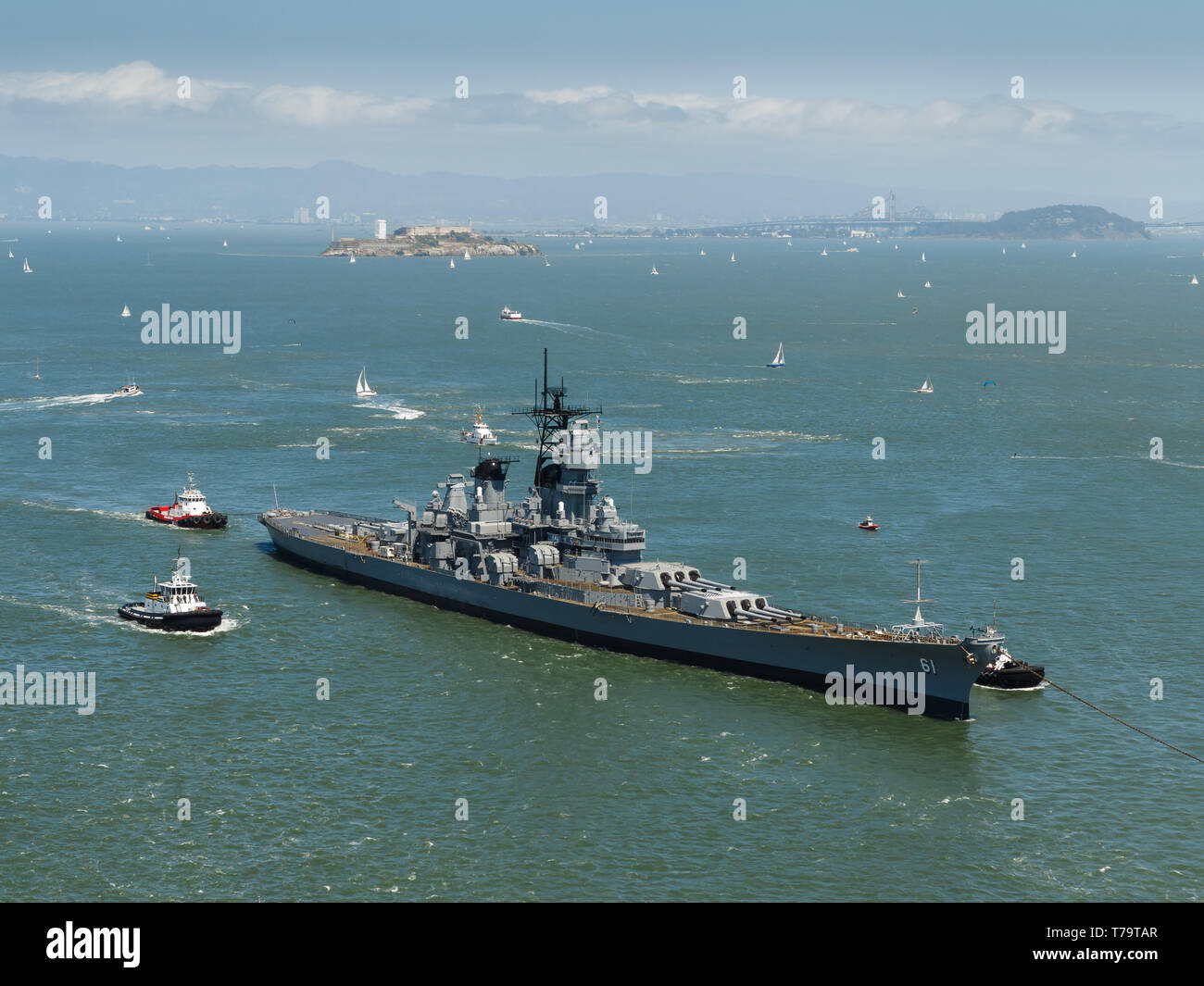 Closeup view of the side of a navy world war II battleship, and tugboats leaving San Francisco harbor with hundreds of sailboats dotting water under s Stock Photo