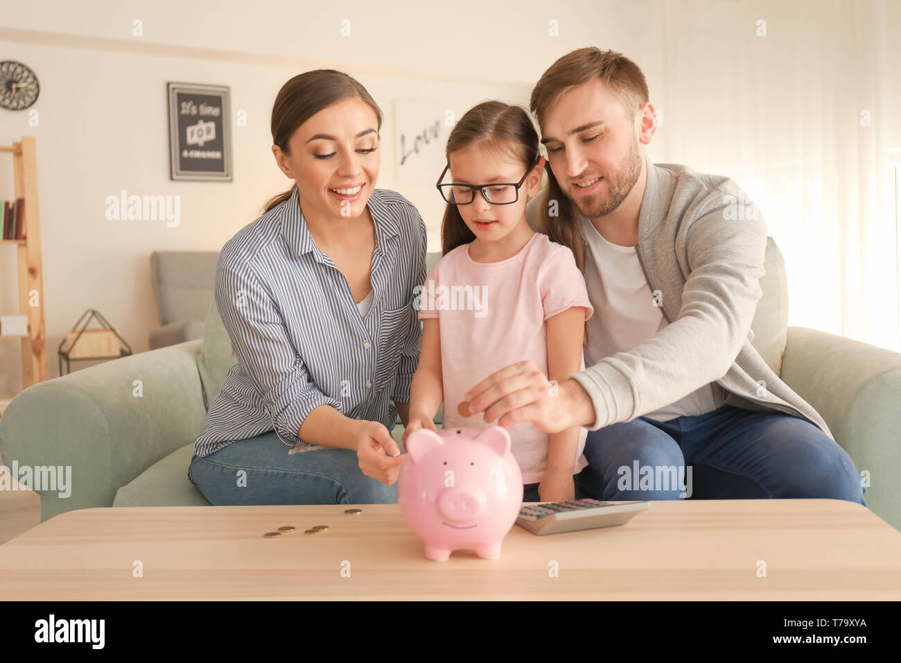 Little girl with her parents putting coins piggy into bank indoors ...