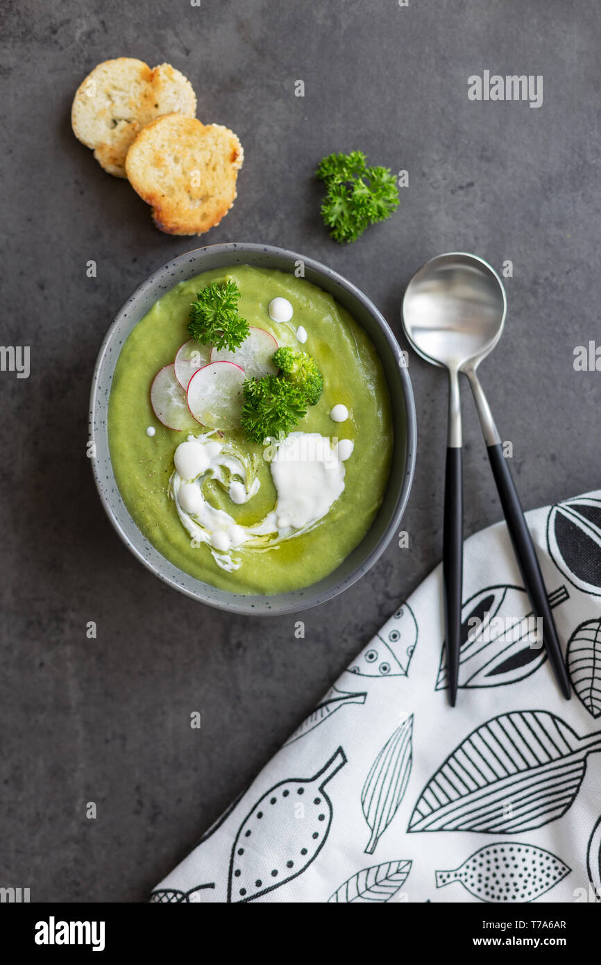 Hot cream soup with broccoli and cream and radish in bowl, bread and spoon at gray background, white napkin near on concrete background. Concept of he Stock Photo