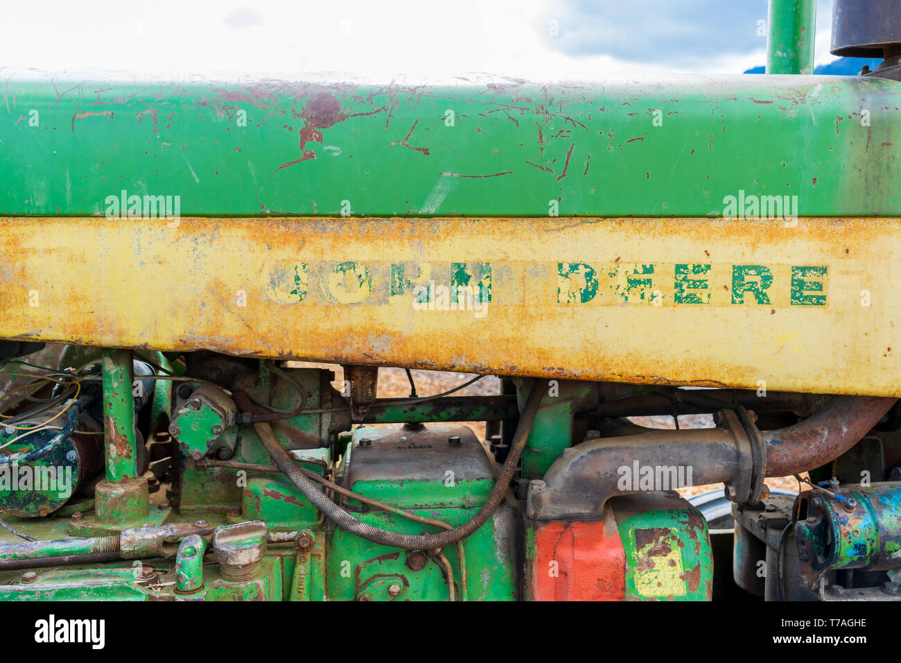 Side of an old, worn down classic tractor, showing remnants of the John Deere logo word mark in green and yellow, and tractor engine parts. Stock Photo