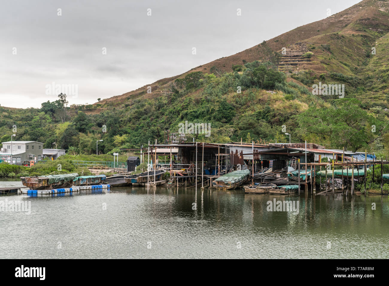 Hong Kong, China - March 7, 2019: Fishermen houses on stilts on Tai O River with large green hill in back under gray sky. Sloops, tarps and simple dwe Stock Photo