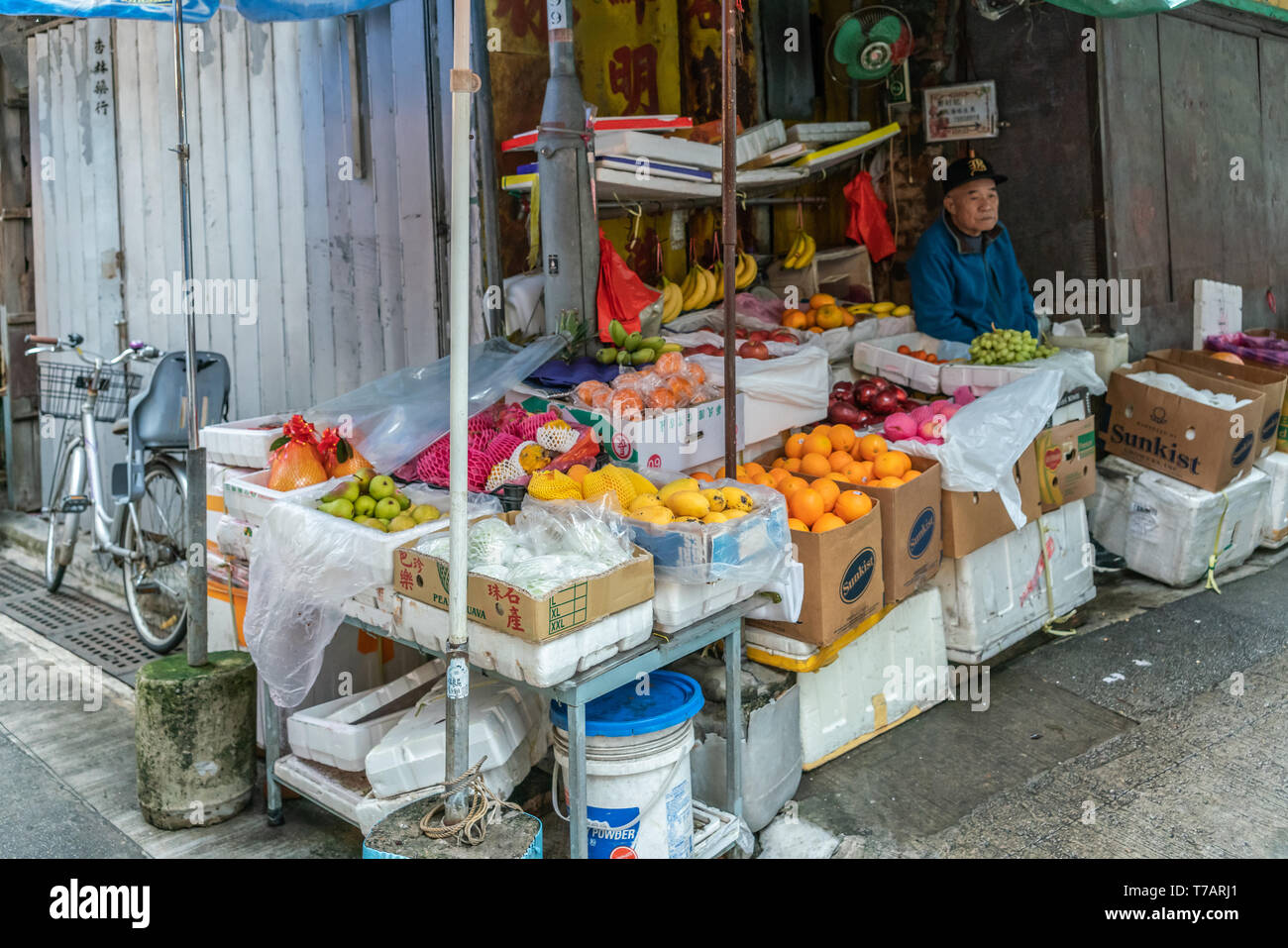Hong Kong, China - March 7, 2019: Tai O Fishing village. Fruit booth with rich choice on corner of street. Older male vendor in blue vest. Colors by t Stock Photo