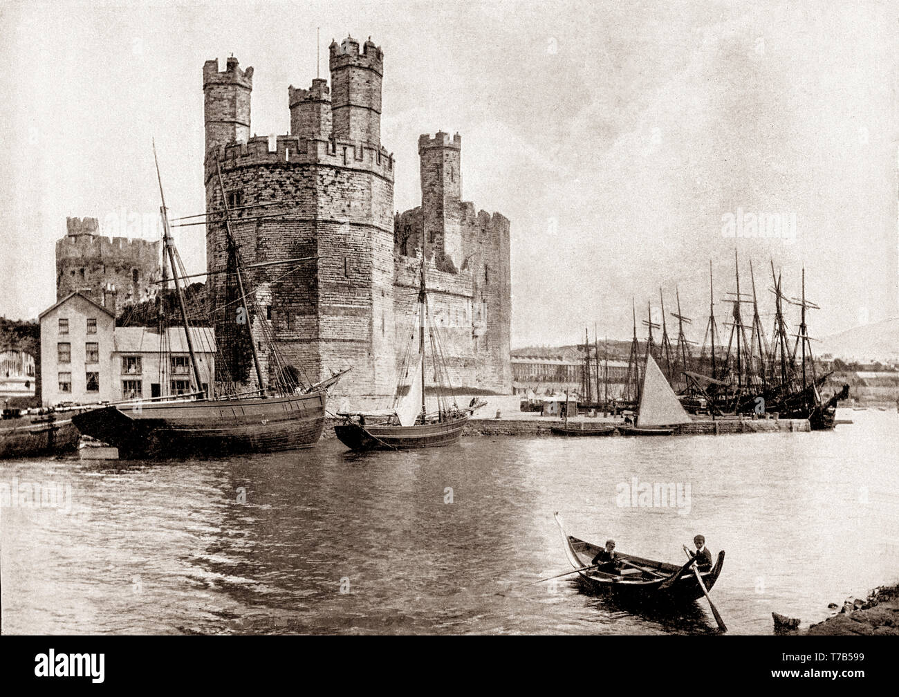 A late 19th Century view of two young boys rowing across the harbour to the  13th Century castle in Caernarfon, royal town and port in Gwynedd, Wales on the eastern shore of the Menai Strait, opposite the Isle of Anglesey. Caernarfon castle, constructed between 1283 and 1330 by order of King Edward I, stands at the mouth of the River Seiont which creates a natural harbour where it flows into the Menai Strait. Stock Photo