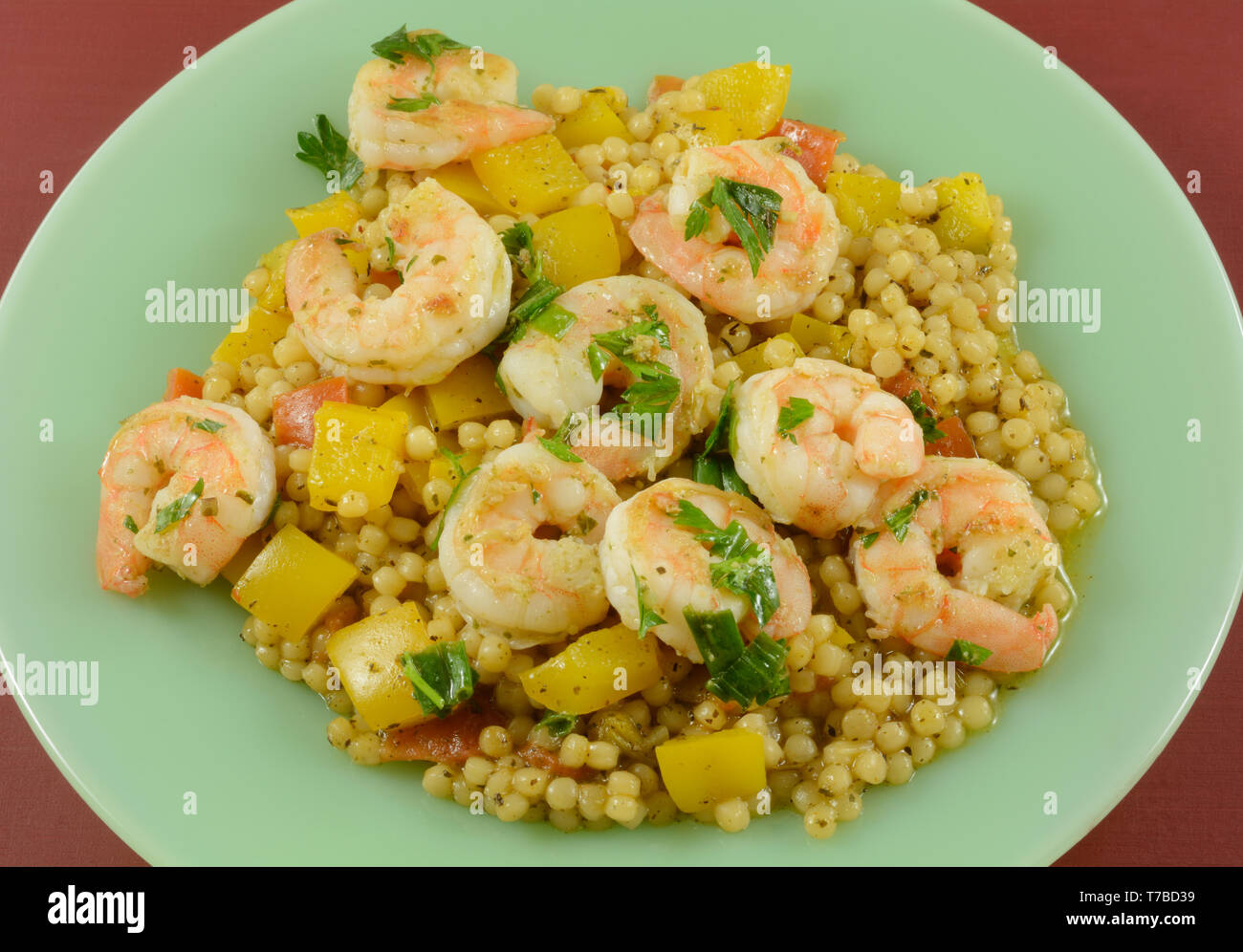 Shrimp, yellow bell peppers and pearl couscous dinner on green plate on red table Stock Photo