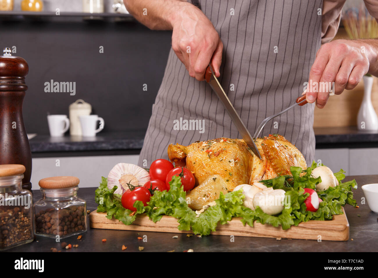 Hands of chief man cuting baked chicken with vegetables with a big knife on a wooden plate in a restaurant Stock Photo