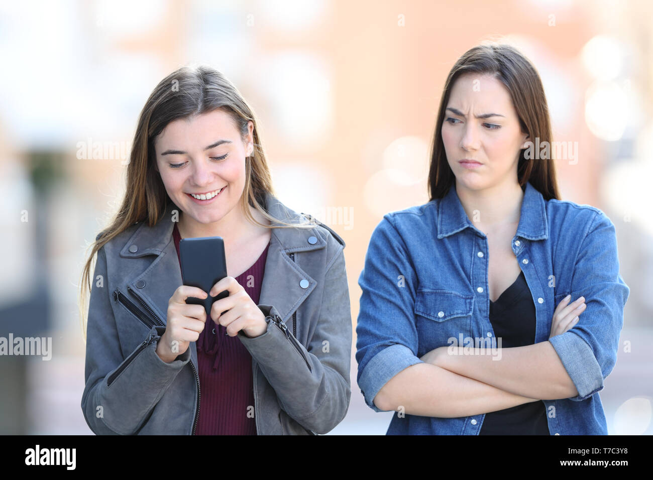 Front view portrait of an angry woman with her friend who is using phone in the street Stock Photo