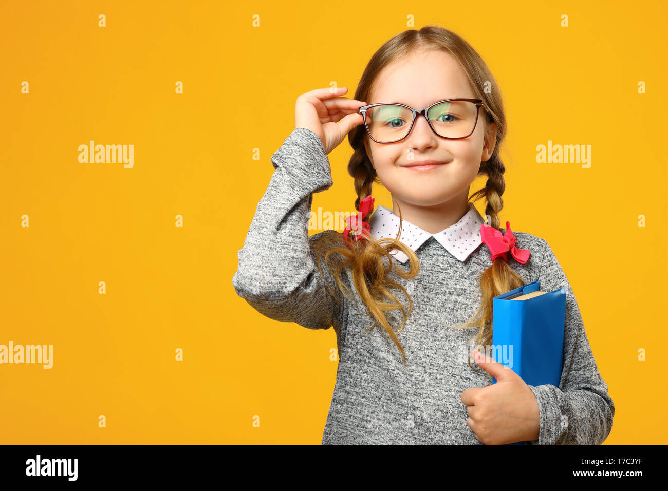 Portrait of a cute little kid girl on a yellow background. Child schoolgirl looking at the camera, holding a book and straightens glasses. The concept Stock Photo