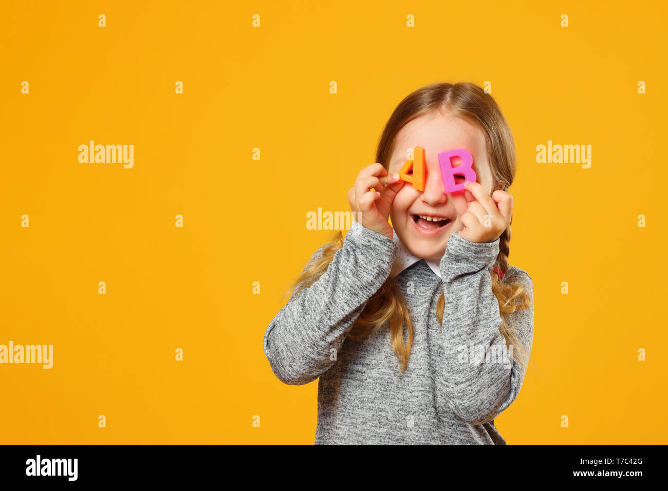 Portrait of a cheerful little girl child on a yellow background. Schoolgirl holds the letters A and B. The concept of education and school. Stock Photo