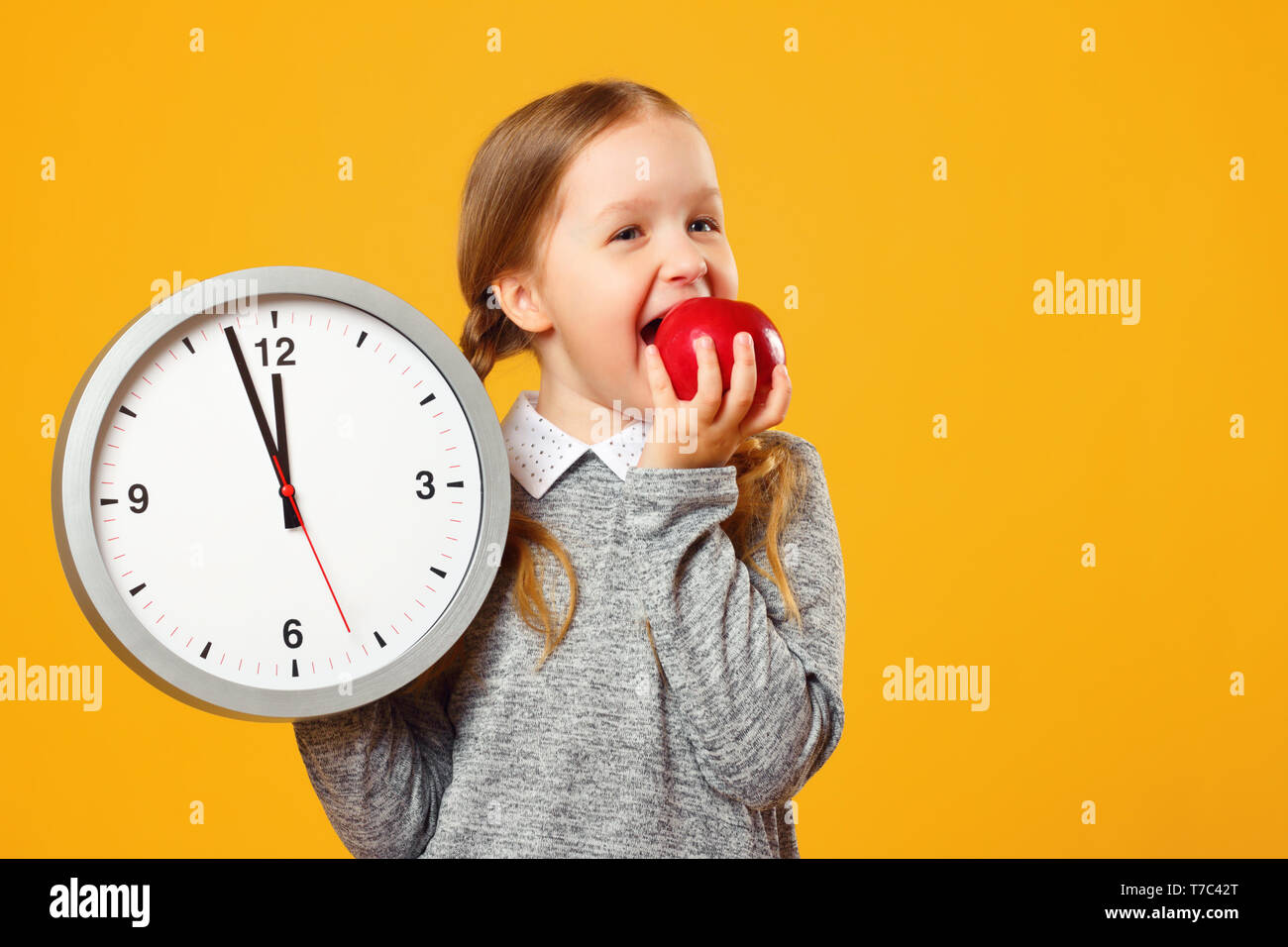 Little girl schoolgirl holds a big clock and bites a red apple on a yellow background. Break and lunch. Stock Photo