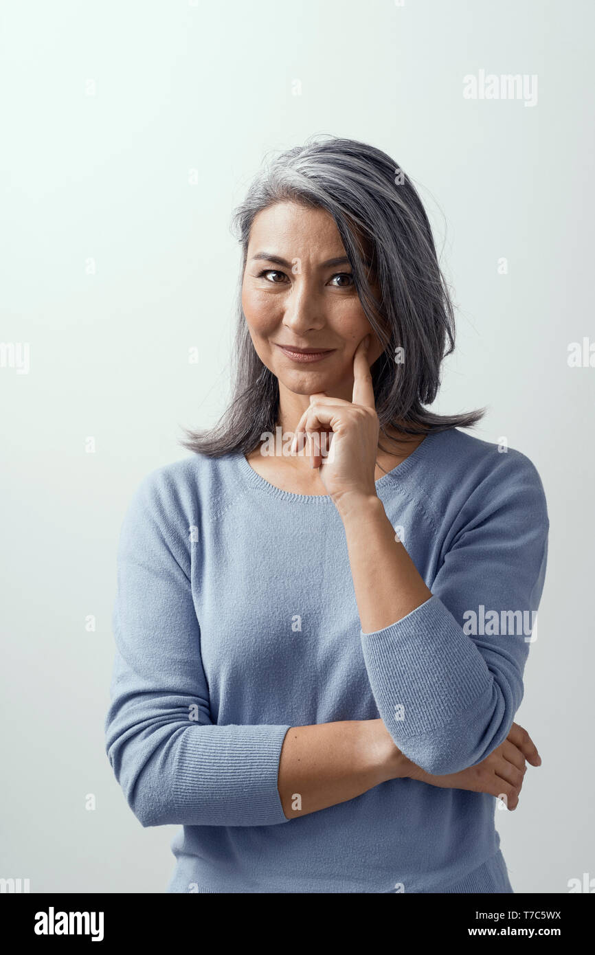 Smiling Asian Woman With Grey Hair Stands And Touches Her Cheek With Pointing Finger Another Hand Is On Chest Charming Middle Aged Lady In Casual Cl Stock Photo Alamy