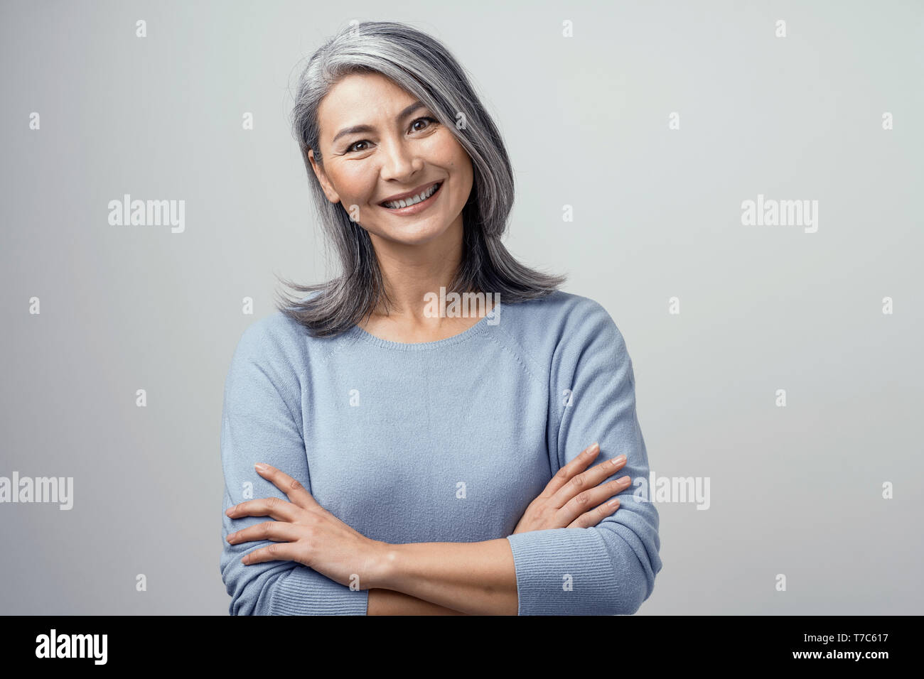 Attractive Optimistic Grey-Haired Mature Asian Woman Smiles Widely In Studio and Crosses Her Hands, Head Tilted. Hands and Shoulders Tonned Portrait O Stock Photo