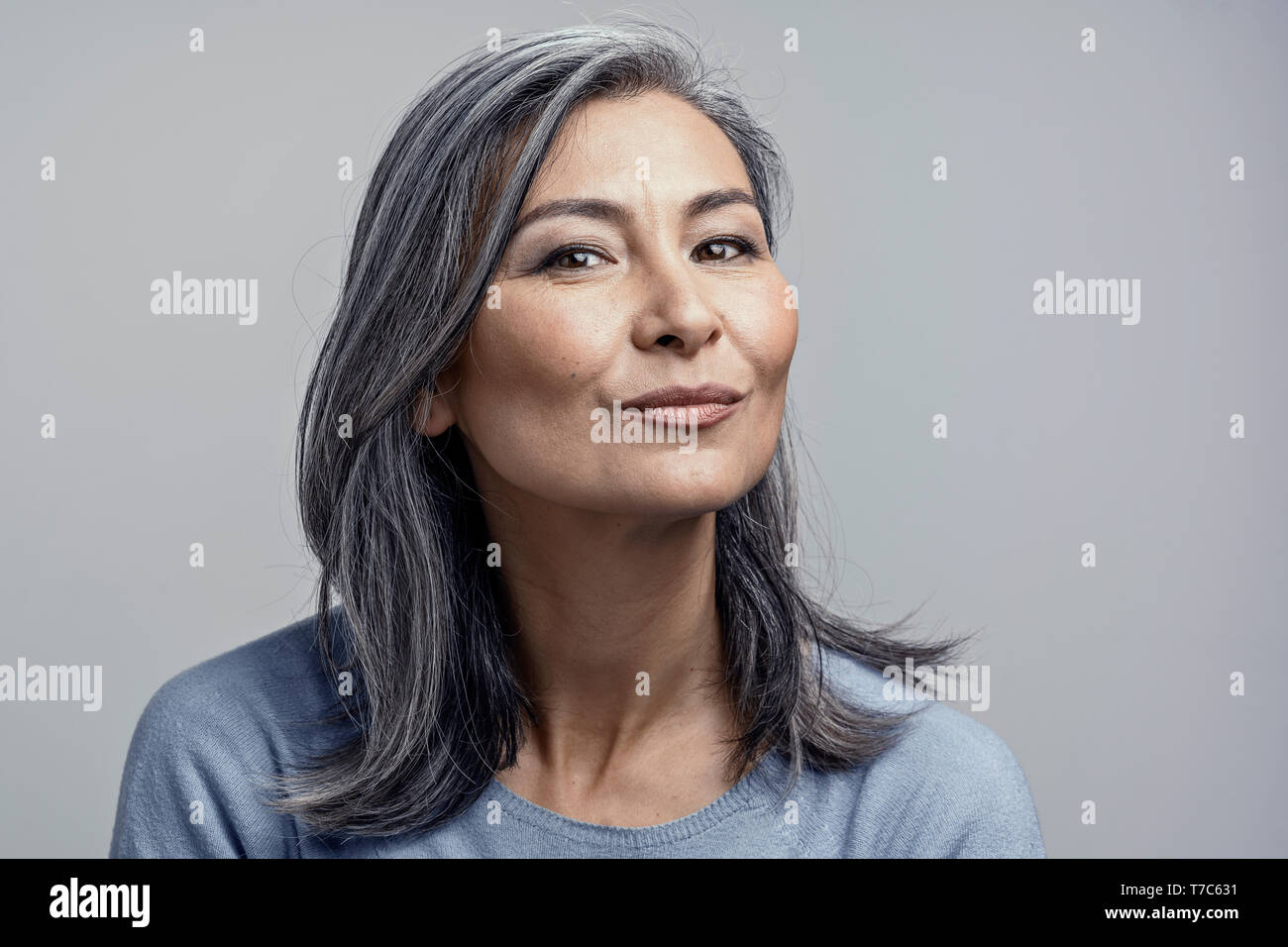 Gorgeous Atrractive Korean Woman with Gray Wavy Hair and Cheekbones Smiles Gently to Camera. Closed-Up Tilted Head and Shoulders Toned Studio Portrait Stock Photo