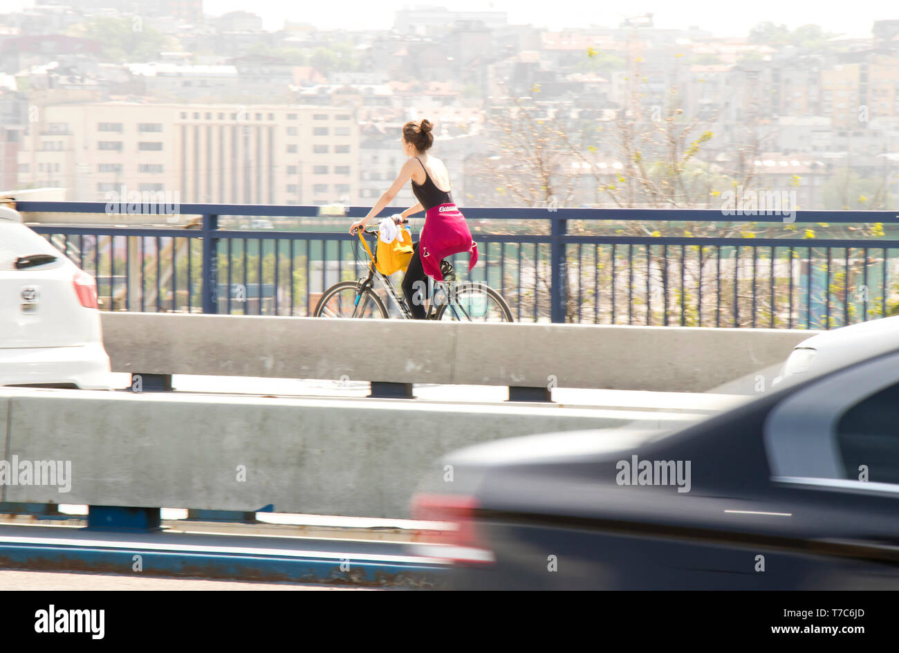 Belgrade, Serbia - April 25, 2019: One young woman riding bike on city street bridge lane with fast traffic in the front and blurry bright cityscape b Stock Photo