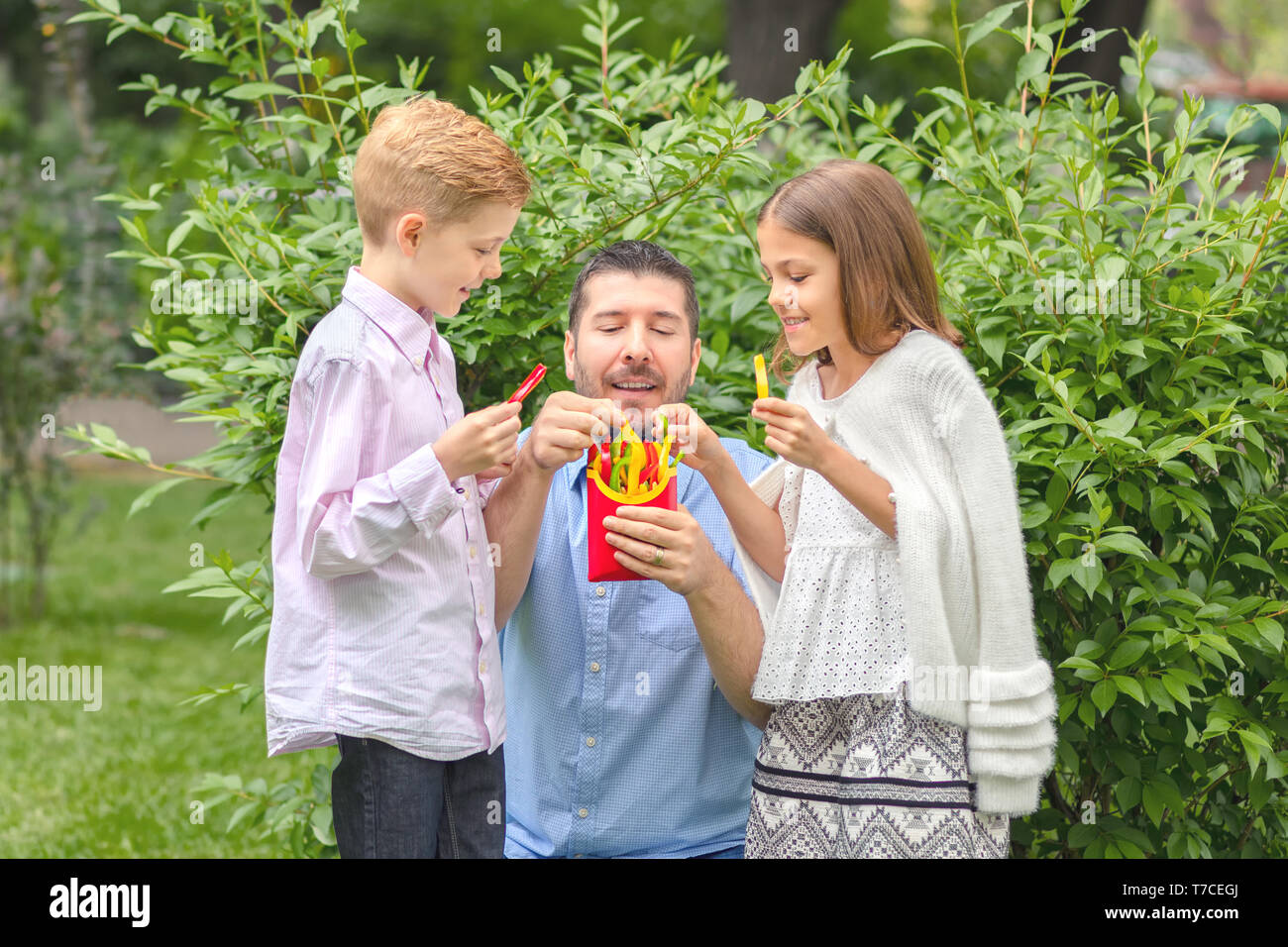 Smiling father and kids eating fresh vegetables in nature – happy dad holding colorful peppers sliced in form of french fries for children Stock Photo