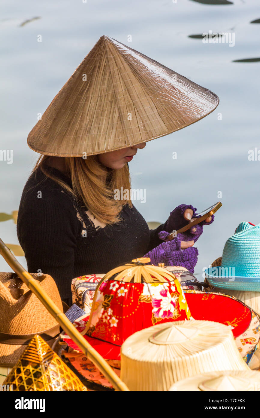 Damnoen Saduak, Thailand - 4th March 2017: Hat vendor on mobile phone.  The floating market is a popular tourist atrraction Stock Photo