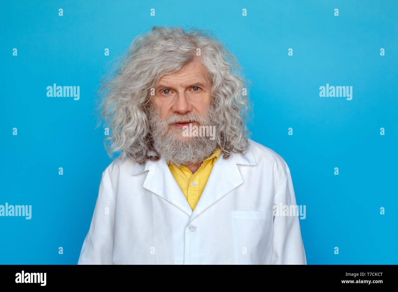 Old long-haired man wearing doctor's gown studio isolated on blue wall looking camera thoughtful Stock Photo