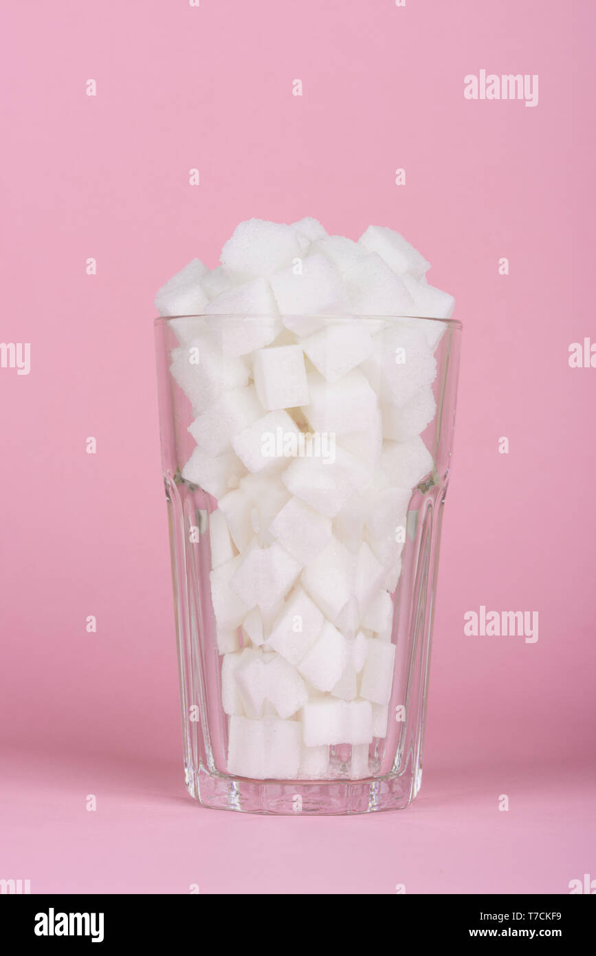Drinking glass of of lump sugar cubes on pink pastel background. Unhealthly diet with sweet sugary soft drinks concept. Stock Photo