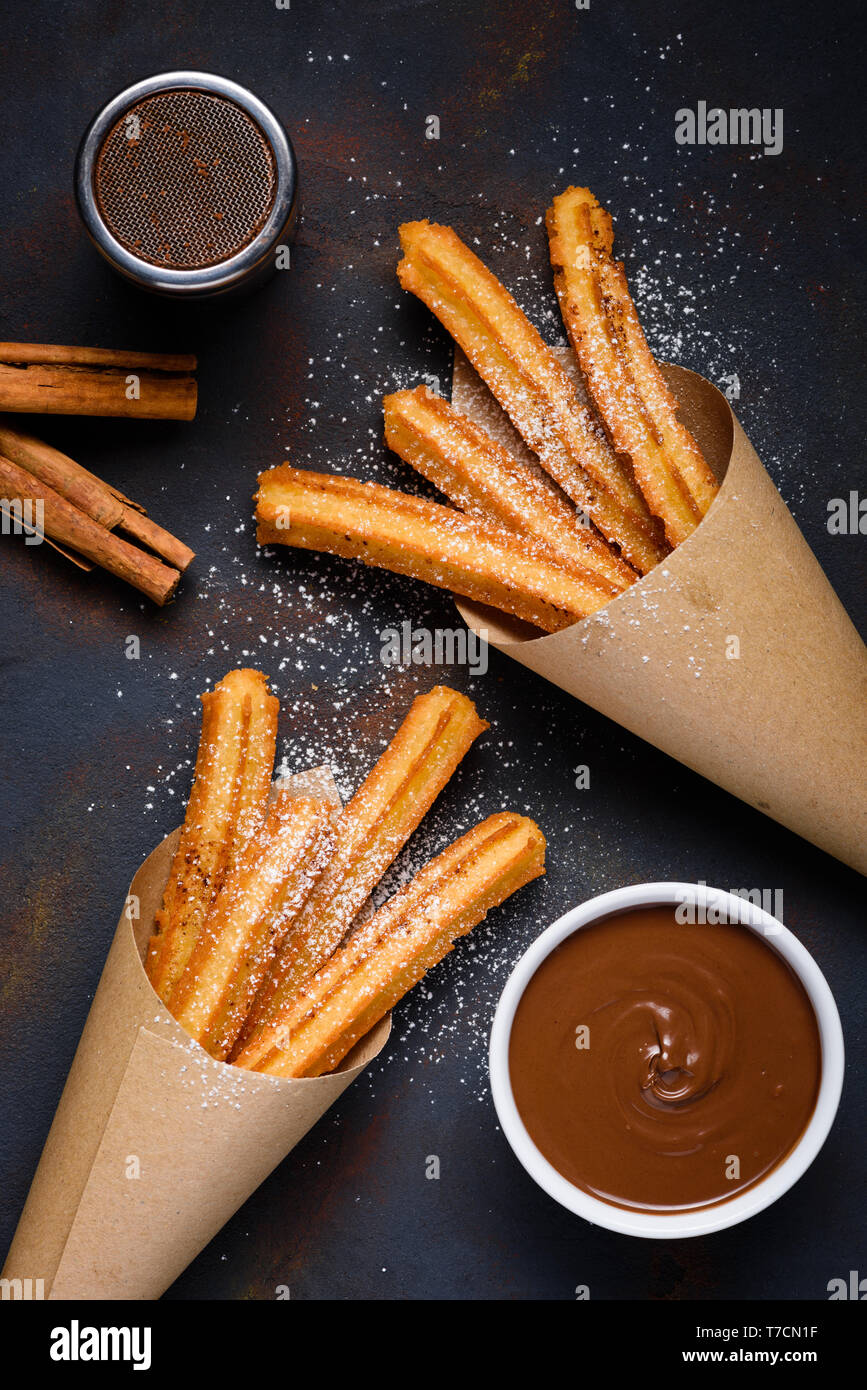 Churros with sugar powder with chocolate sauce dip and cinnamon sticks on dark background top view Stock Photo