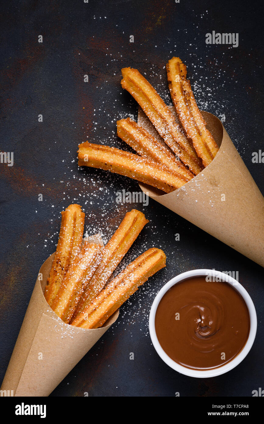 Traditional churros sticks in paper bag with sugar powder cinnamon and bowl of chocolate dip on dark table top view Stock Photo