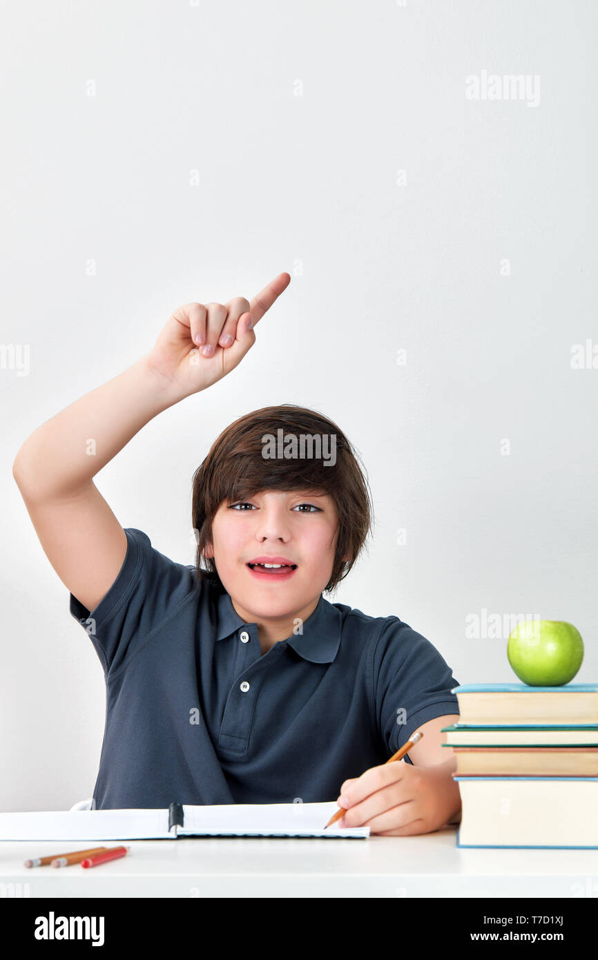 Excited and happy smiling caucasian boy sitting at the desk raising hand with index finger to answer the question in classroom at school Stock Photo