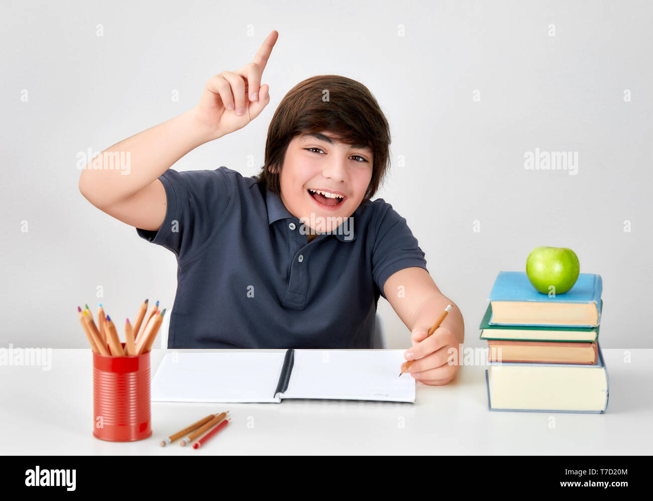 Excited and happy smiling caucasian boy sitting at the desk raising hand with index finger to answer the question in classroom at school Stock Photo