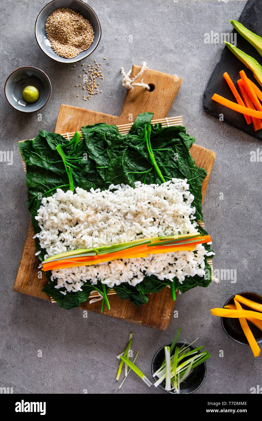 Springlike vegan maki sushi in the making with spinach, avocado, carrots, mango and shallots on wooden board over grey concrete background Stock Photo